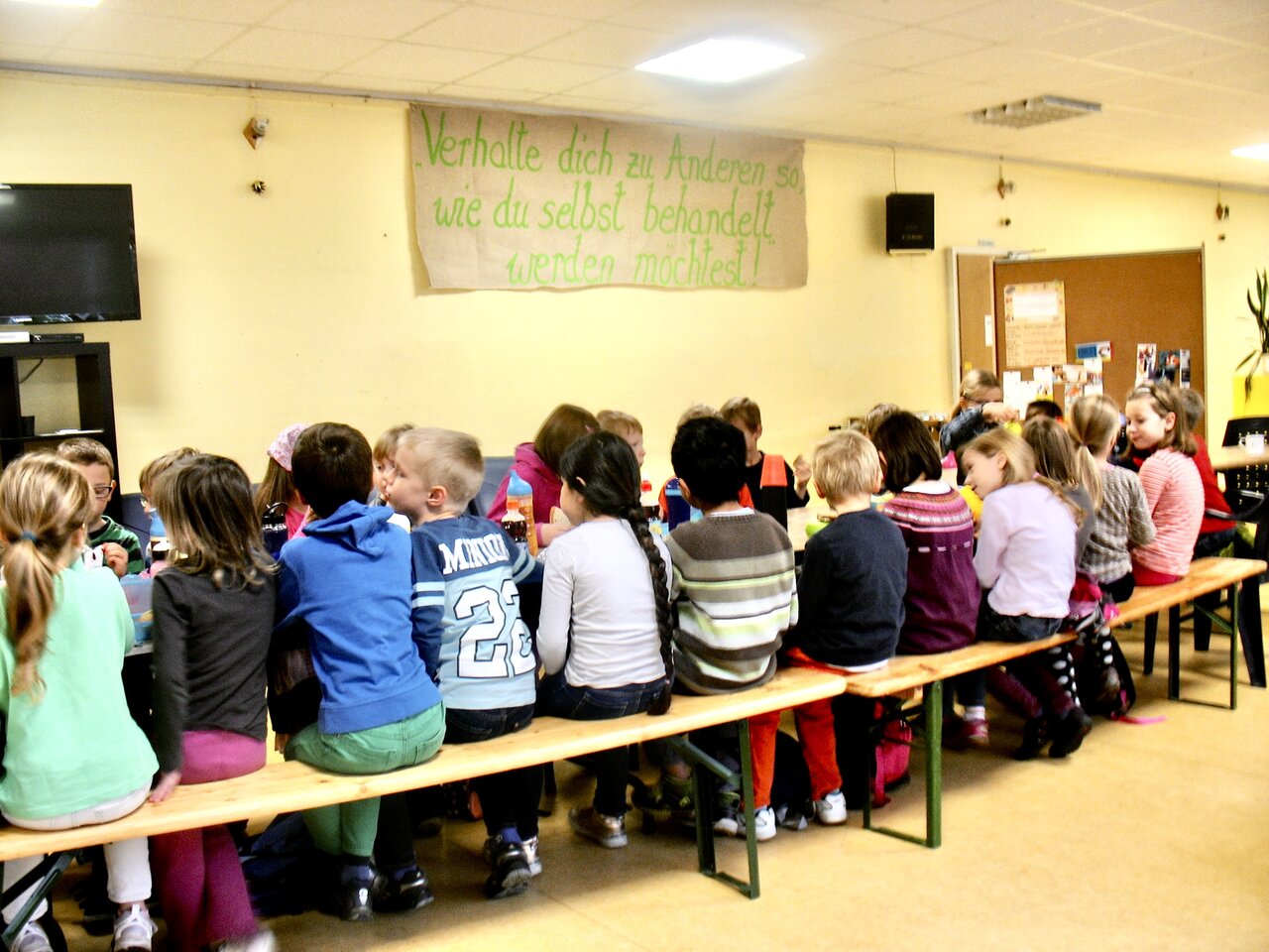 Eine große Gruppe von Kindern mit dem Rücken zur kamera. Man sieht sie an einem langen tisch im Kinderclub sitzen im Rahmen eines Fairnesstages, einer damaligen Veranstaltung für Schulklassen