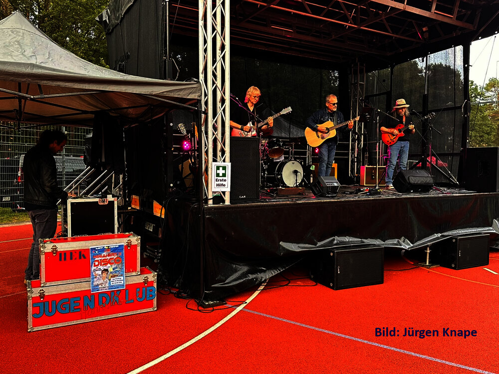 Eine Bühne auf dem roten Sportplatz beim Stadtteilfest. Darauf eine vierköpfige Band, daneben ein Techniker an einem Mischpult