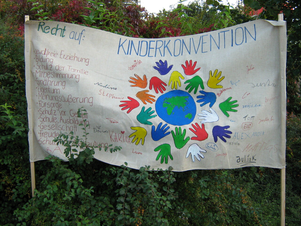 Ein weißes Banner mit einer gemalten Weltkugel und Kinderhänden, daneben gelistet die Rechte von Kindern, deren Vernkerung im Grundgesetz gefordert wurde, z.B gewaltfreie Erziehung und Bildung