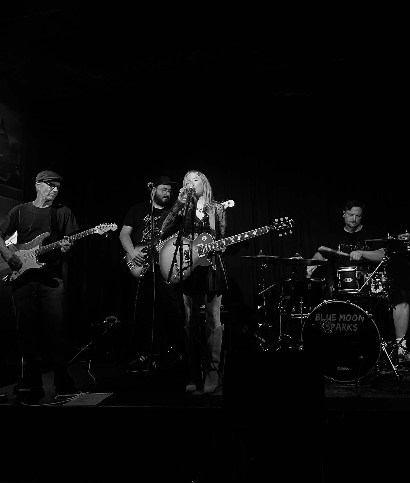 schwarz-weiss Bild mit der Band vor schwarzem Hintergrund, eine Frau mit E-Gitarre steht am Mikrofon, dahinter Männer mit Gitarre, Bass und Schlagzeug